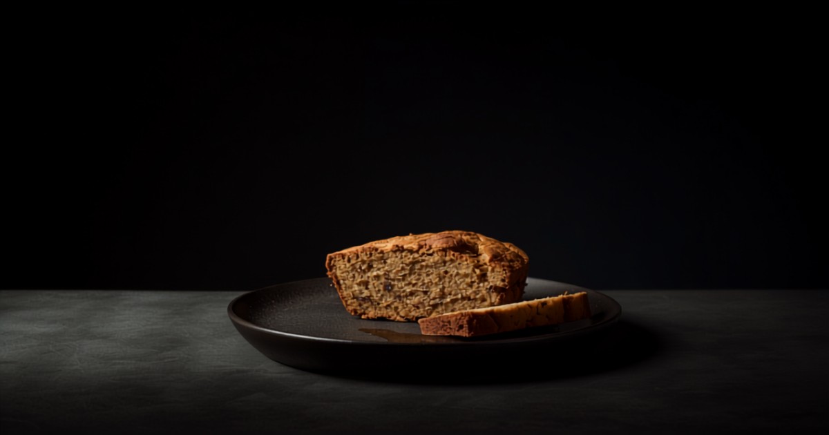 Pan de plátano cortado mostrando la miga, con una rebanada sobre cerámica oscura en penumbra ocre.